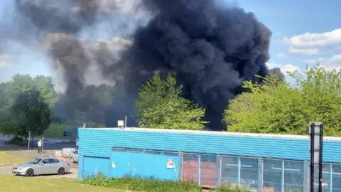 Paul Yates Large plume of dark smoke rises above a blue warehouse with trees in the foreground