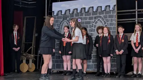 A teacher directs two teenagers on stage in their scene. The ensemble stand behind them along the back of the stage. All the cast are in school uniform.