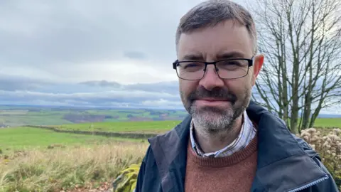 Dave Richardson is a middle-aged man with short hair, a trimmed beard and glasses. He is wearing a brown jumper and a blue coat and behind him is a vista of rolling fields and a grey sky