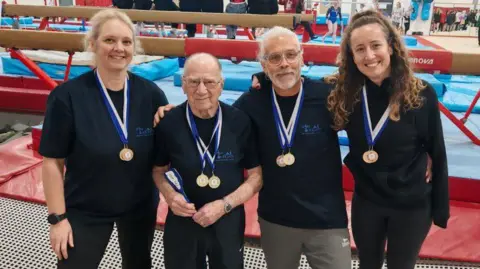 Sue Burt Quinney stands second from the left, with two female competitors each end and another man to the right in the picture. They are all wearing medals and smiling at the camera wearing navy t shirts. Behind them are gymnastics beams and they are standing on a trampoline.
