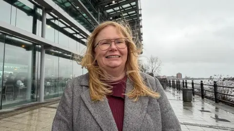 Carolyn Basing is wearing a grey woollen coat and claret jumper and standing in front of the National Glass Centre. Through the windows people can be seen eating in the cafe. She has long dark blonde hair which is being moved by the wind. It appears to have been raining and the River Wear can be seen on the right under a grey sky.