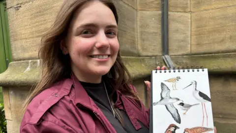 BBC/Simon Thake A smiley woman with dark hair and a purple jacket holds up a book of hand drawn pictures of birds.