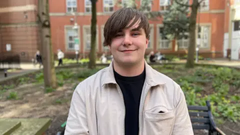 Covurn is wearing a black t shirt and a cream jacket. He is in a park in Leeds