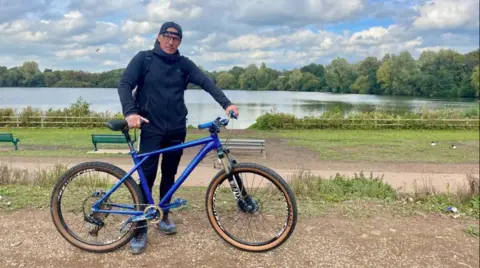 A man in standing beside a lake, holding a blue bicycle. He is wearing a dark zipped hoodie, a black rucksack, glasses and a dark baseball cap worn backwards. His left hand is holding the handlebar of a bicycle. Geese and green benches can be seen in the background beside a footpath.