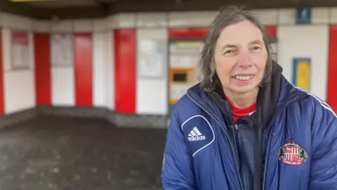 Paula Mitchinson is smiling against the red and white backdrop of the Stadium of Light metro station. She is wearing a blue Adidas puffer jacket with Sunderland AFC's crest and has short grey hair.