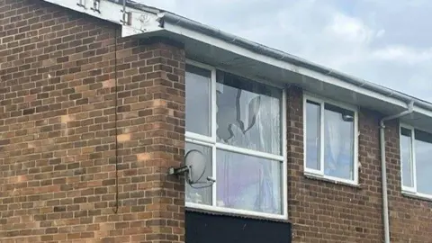 Sam Smith A large smashed window of an upstairs flat. The modern flat building is made of brown brick; the pane of glass in a large window at the end of the property has been badly broken.
