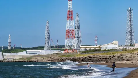 Two people walking on a beach, with the Kashiwazaki-Kariwa nuclear power plant in the background.