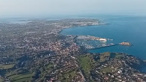 BBC An aerial shot of Guernsey showing a harbour and a bay with boats, buildings, green spaces and trees with a blue sky.