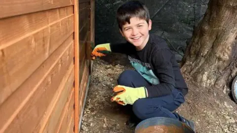 Family handout/PA Real Life Harry is seen crouched down painting a wooden fence. A tub of brown paint is seen beside him as he is at the edge of a garden with a tree behind him.