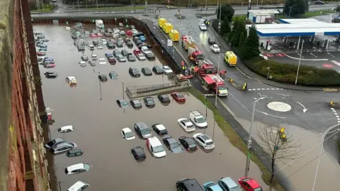 Taken from a top-storey window, a large carpark is full of brown floodwater, which reaches the windows of the cars. Rescue vehicles are parked on a sloped road above.