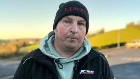 BBC Jonathan Potter is looking directly at the camera. He has pursed lips and is wearing a black beanie hat, a grey hoodie and a black jacket. There are green fields in the background.