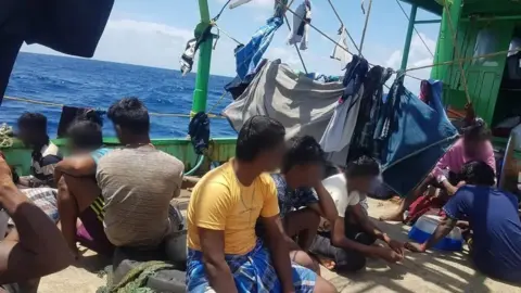 Selebaran Para migran Tamil, dengan wajah kabur atau memalingkan muka dari kamera, duduk di dek kapal, dengan tali jemuran di sekeliling mereka, saat mereka berlayar melintasi Samudera Hindia