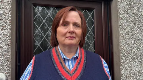 A middle-aged woman is standing in front of the door of a house. The door is dark brown wood effect, with a leaded style glass panel. She has chin length auburn hair and is wearing a blue knitted vest top over a blue and white striped shirt. 