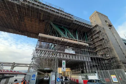 LDRS The Tyne Bridge is partially covered by scaffolding around one of its towers. There is a road and pavement with cars and workers.