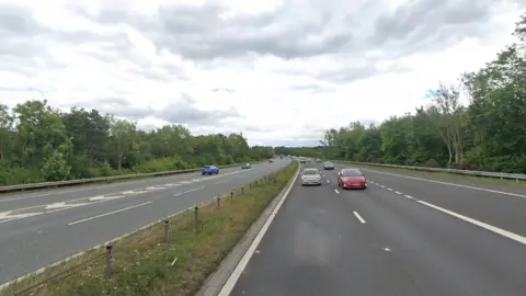 The motorway has six lanes with a grass strip and a small fence down the middle. There are about a dozen cars on the rod. The road is bordered by tall green trees.