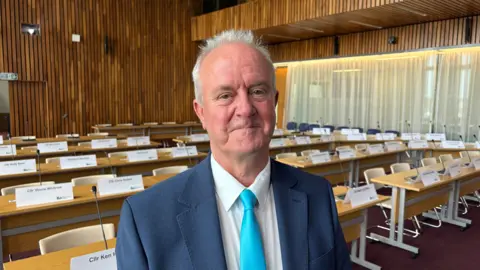Martin Griffiths with short white hair, smiling at the camera and wearing a white shirt, light blue tie and dark blue jacket. He is standing in front of rows of wooden desks with chairs behind. There are white name labels in front of each chair. There is a wood panelled wall at the back of the room, and windows with thin curtains on the right.