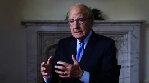 George Mitchell. He has grey hair at the sides, wearing glasses, a navy blazer, light blue shirt and blue tie. He is holding his hand out to the front. A marble fireplace is behind him.