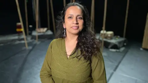 Ant Saddington/BBC Urja Desai Thakore in a green outfit on a stage smiling into the camera. She has long, curly black hair and long earrings.