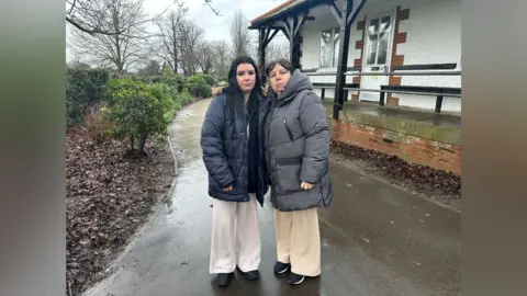 A woman and her daughter. They are wearing big winter coats. Both look serious. 