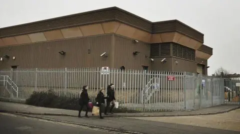 Getty Images A security fence surrounds the Securitas depot in February 2006 in Tonbridge. The flat-roofed building has cameras around it. Pedestrians are walking past the site. The warehouse, which has brown cladding, is photographed under a grey sky.