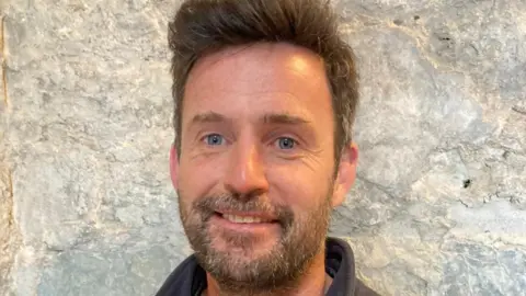 Ryan Byrne has blue eyes, brown hair and a beard.
He is wearing a grey fleece and standing in front of a stone wall.