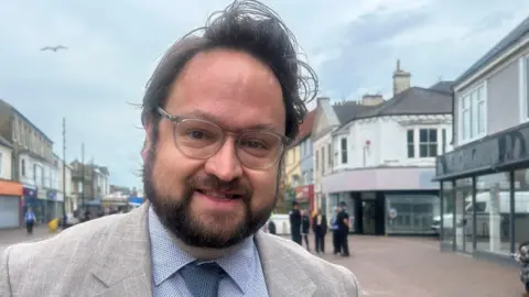 Matt Storey is standing on a high street and smiling slightly at the camera. He has short dark hair and facial hair and is wearing glasses and a grey suit. There are police officers speaking with members of the public behind him, but they are out of focus.