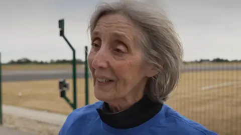 An elderly woman wearing a blue shirt standing beside a fence near an airfield runway.