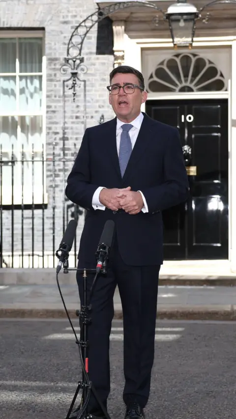 Andy Burnham stands in front of a microphone outside the black door of 10 Downing Street, wearing a dark suit and light blue tie. 