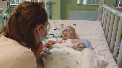 Emily Bowen A newborn baby lying in a hospital cot with various tubes and wires attached to her and a blanket over her. A woman with long brown hair, wearing a face mask, is leaning over her.