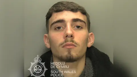 A custody image of a man looking at the camera. He has dark brown hair and a dark stubbly beard and moustache.