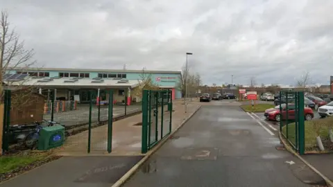 The outside of Bourne Elsea Park Primary Academy. It shows a road leading to a car park. There are green gates which are open. The sky is overcast.