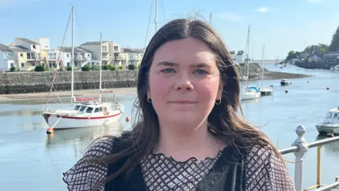 Mared Llywelyn is facing the camera. She has dark hair and is wearing a black dress. Behind her is the Menai Strait. It is a sunny day and there are boats moored in the background.