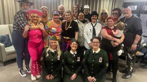 Montagu Hall Care Home A group of 18 women and men and one young child, in an array of fancy dress costumes lined up in three rows smiling at the camera. The front three women are kneeling and are wearing army uniforms costumes