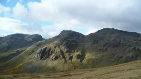 Michael Graham/Geograph Three crags of grey rock and green mossy ground are visible. The middle one is Esk Buttress. In the foreground there is a relatively flat grey-green grassy surface and in the background there is blue sky with some white clouds.