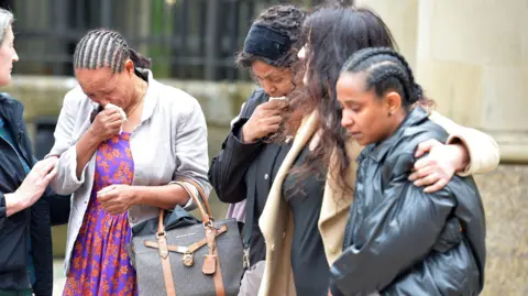 Spindrift A group of women huddled together outside a court building. Two have their heads bowed and are weeping into handkerchiefs.