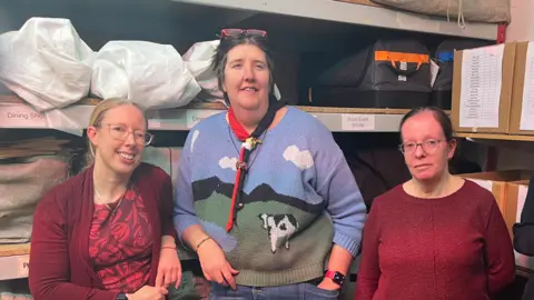 Wayne Bavin/BBC A woman with blonde hair and glasses, a taller woman with dark hair wearing glasses on her head, and a third woman wearing glasses wearing a red jumper standing in front of shelving