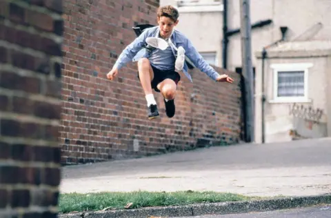 Alamy Character Billy Elliot jumping in the air while holding ballet shoes tied around his neck, against an urban backdrop