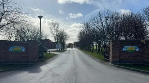 Ellis Bros Group The entrance to Skegness Water Leisure Park has a tree-lined driveway. There are two black metal gates, which are open. There are brown brick walls with the leisure park logo, on both sides. The logo is blue, orange and green.