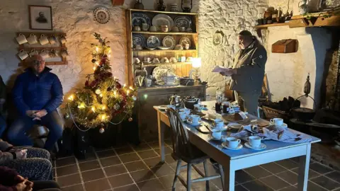 Inside of an old cottage, a kitchen table sits upon a brown tiled floor. It is covered in teacups and the remnants of bread and butter that was supped on.
A short Christmas tree sits on the left glowing, with light and ornaments shining. In the middle, an old fashioned kitchen dresser, filled with fine china and a bright warm lamp.
To the right, Davy Moore stands with glasses on, reading his own poetry to the group on the left.