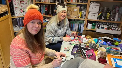 Tracy Wilkinson (left) in an orange hat and beige and orange jumper painting a rock alongside Helen Nelson with long blonde hair, wearing a reindeer hat and grey Christmas jumper. They are smiling.