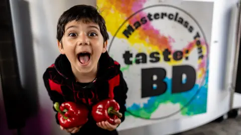 A young boy stands holding some red capsicum peppers and poses joyfully into the camera. He stands in from of a sign that says Masterclass Tent - Taste: BD