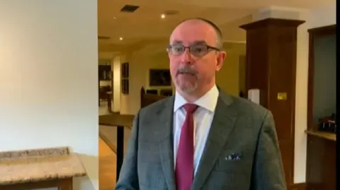Andrew Hollett with short dark hair with a slight beard and round glasses wearing a grey jacket, red tie and white shirt. He is standing in a hotel- a wood pillar is visible behind him to the right and a bar to the left.