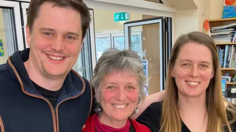 Joy Roodenburg-Vermaat with her son and daughter. Her son has short dark hair and is wearing a navy blue fleece. Joy is stood next to him. She has short grey hair and is wearing a red top. Her daughter is to her right. She has long dark blonde hair and is wearing a black top. All of them are looking at the camera and smiling 