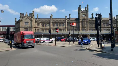 Shrewsbury railway station is in the background, it is a large brown building with a clock tower and dozens of high chimneys. There is a car park in front of it and a road running parallel to it. A red lorry is driving on the road, through some traffic lights.