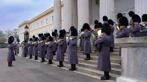 Contributed Captain Claire Lawrence, conducting a group of musicians on steps at Sandhurst. They are all wearing bearskin hats and grey coats and trousers, with black shoes. They are all playing musical instruments. 