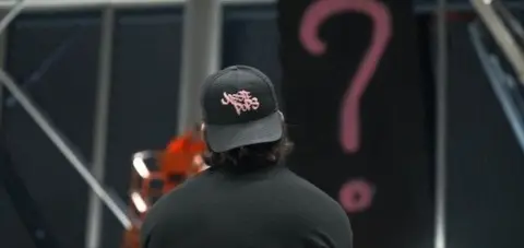 Dillon Osborne The back of a man with shoulder length black hair, wearing a black top and a black baseball hat, worn backwards with pink writing saying Jossiepops on it.
He appears to be looking a painting, covered in a black covering with a pink question mark on it.