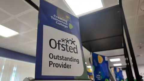 Creating Tomorrow College A blue and white banner with the words 'Ofsted Outstanding Provider' and the Creating Tomorrow College logo. In the background, a school room with overhead lighting can be seen. 