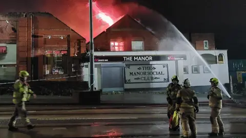 South Yorkshire Fire & Rescue Four firefighters on the road outside a building which has a sign saying Manor Social Club on the front. A cordon can be seen tied to one side of the building. A jet of water is aimed at a large red flame within the building.