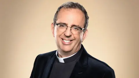 BBC/Ray Burmiston Rev Richard Coles wears a clerical collar and black blazer while smiling directly into the camera.