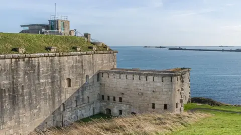 Ian Capper View from Nothe Fort looking out to sea with the Portland Harbour walls in the distance. In the foreground is a section of the Victorian stone fort with a more recent concrete structure built on the top.